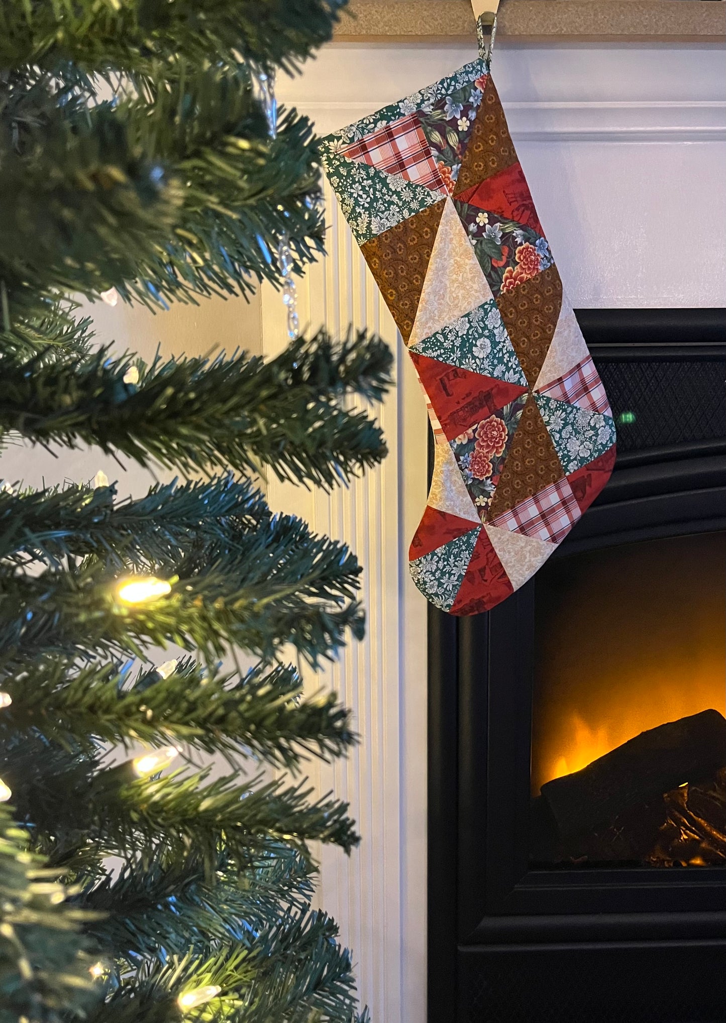Quilted patchwork Christmas stocking hanging by a fireplace with a lit Christmas tree.